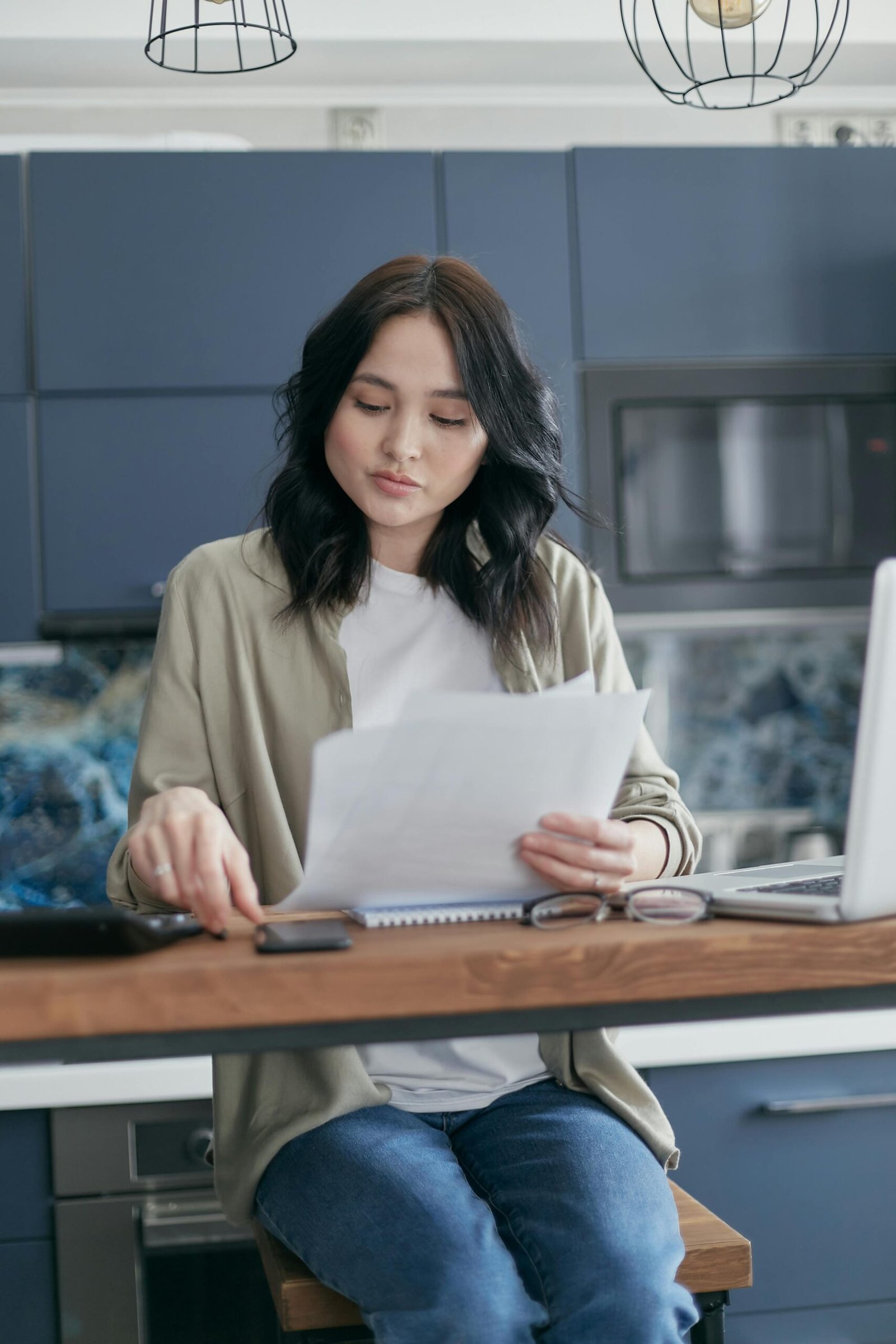A young woman reviews documents and uses her laptop in a modern kitchen setting.