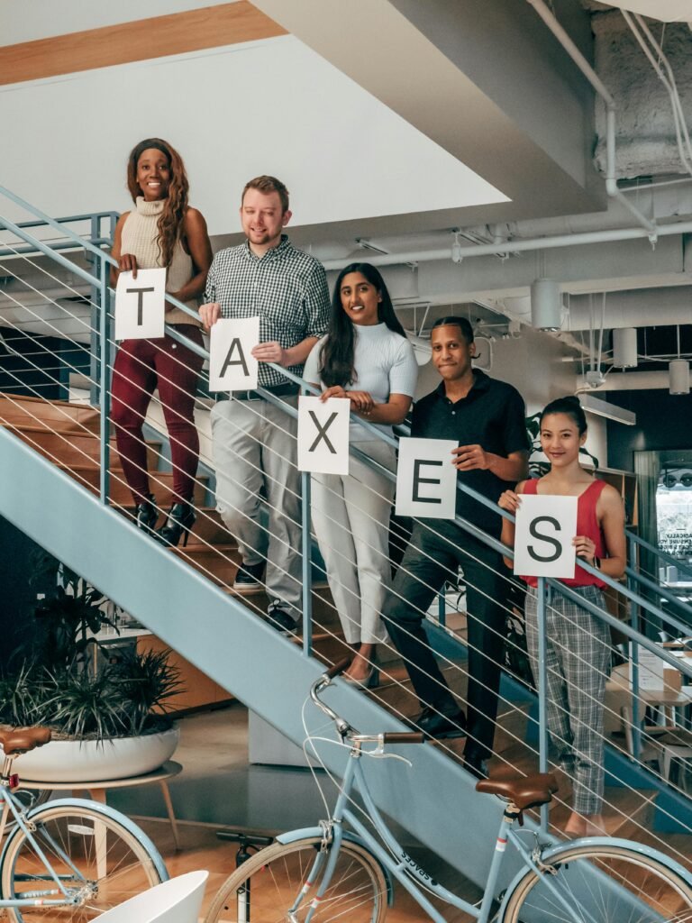 A group of diverse colleagues holding 'TAXES' sign while standing on a modern office staircase.